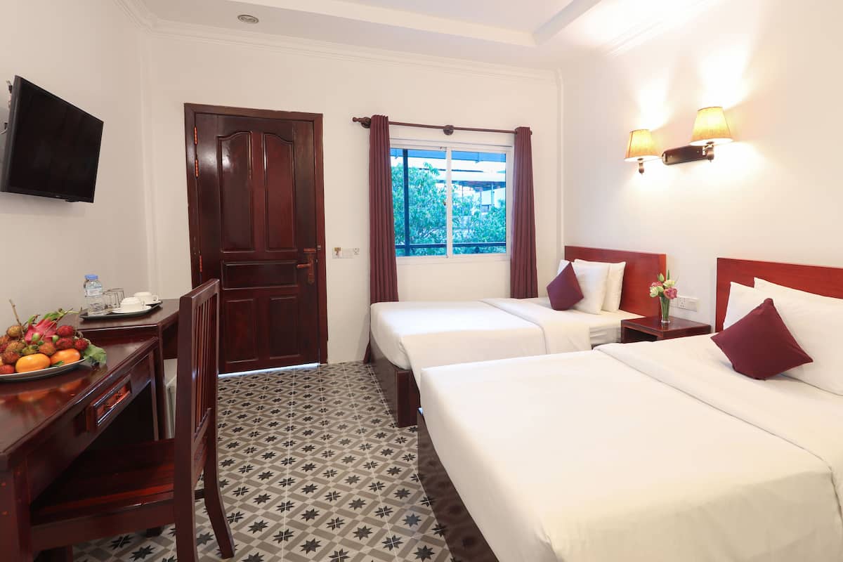 A hotel room with two beds, a desk with a fruit basket, a TV, and a window with curtains.