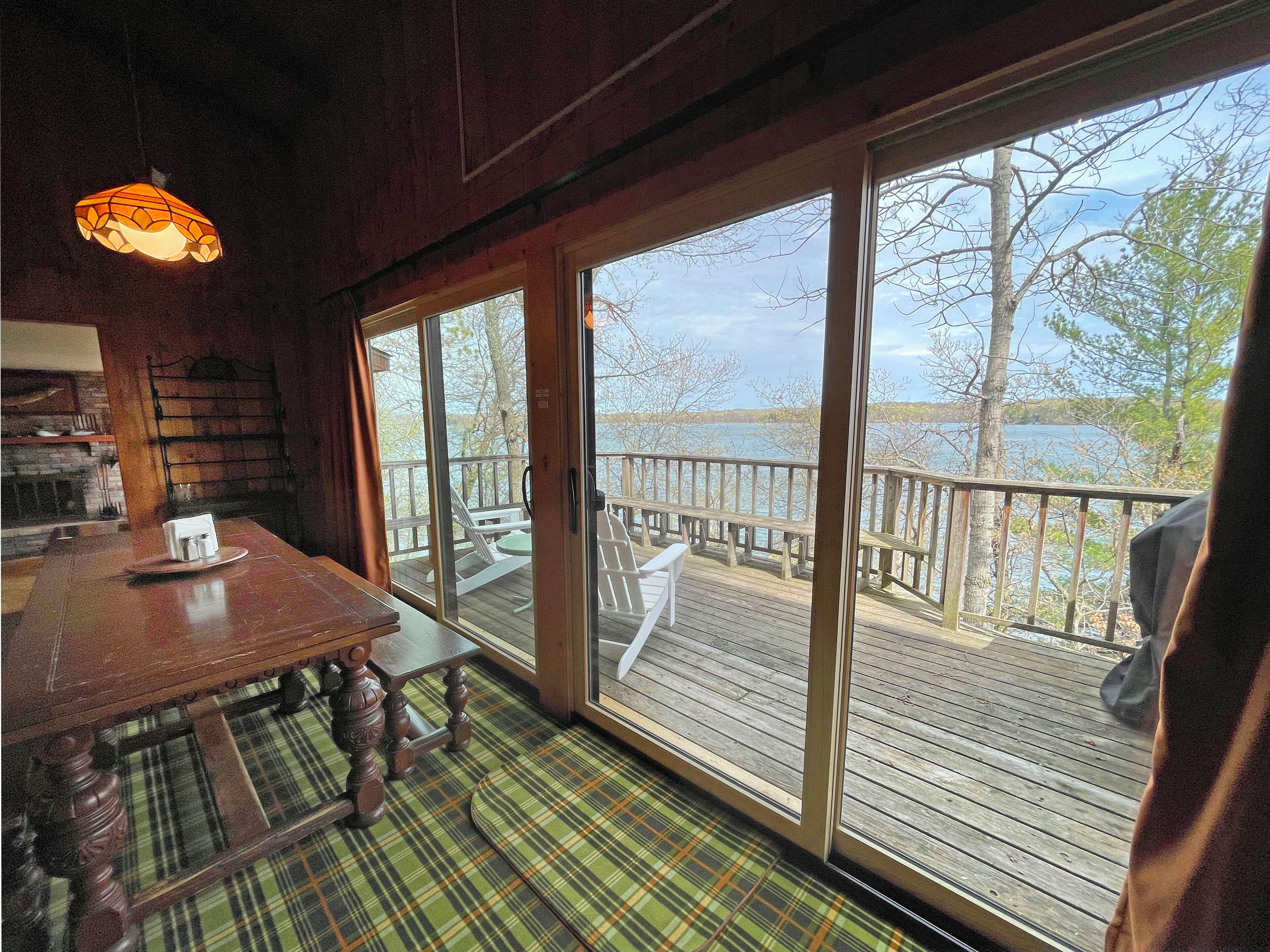 Un intérieur en bois comprenant une table et des chaises, un tapis à motifs écossais et une grande fenêtre donnant sur une terrasse en bois et un plan d’eau.