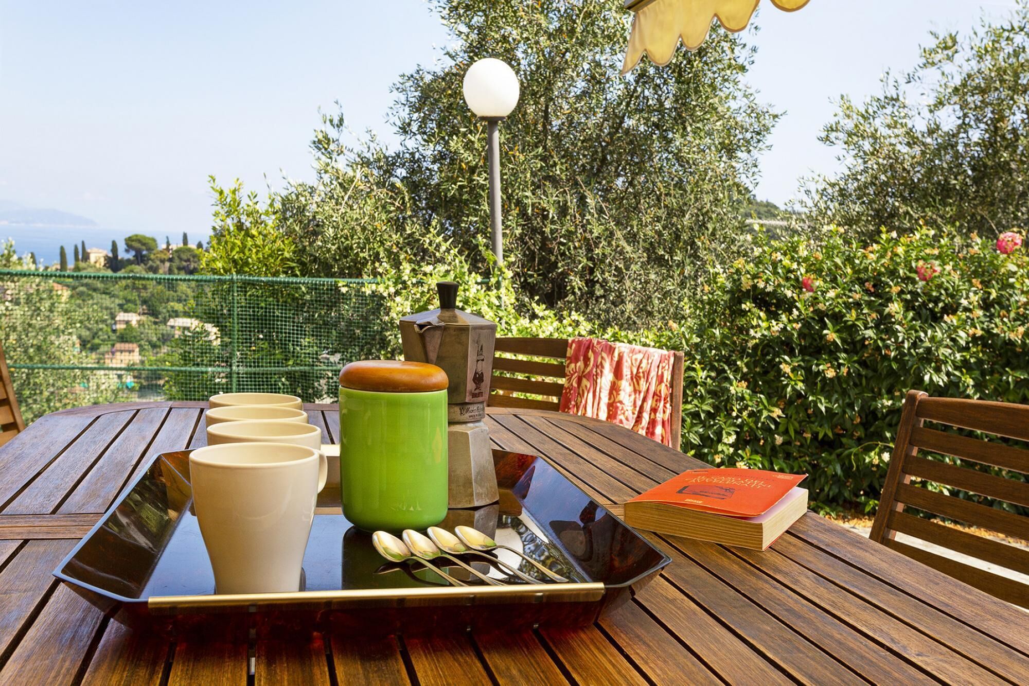 Une table en bois sur laquelle se trouve un plateau contenant une tasse verte, une tasse blanche et un livre, le tout placé devant un décor de verdure luxuriante et d’un ciel dégagé.
