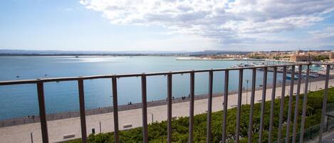 Un paisaje urbano costero con un puerto deportivo, un paseo marítimo y una cadena montañosa distante.