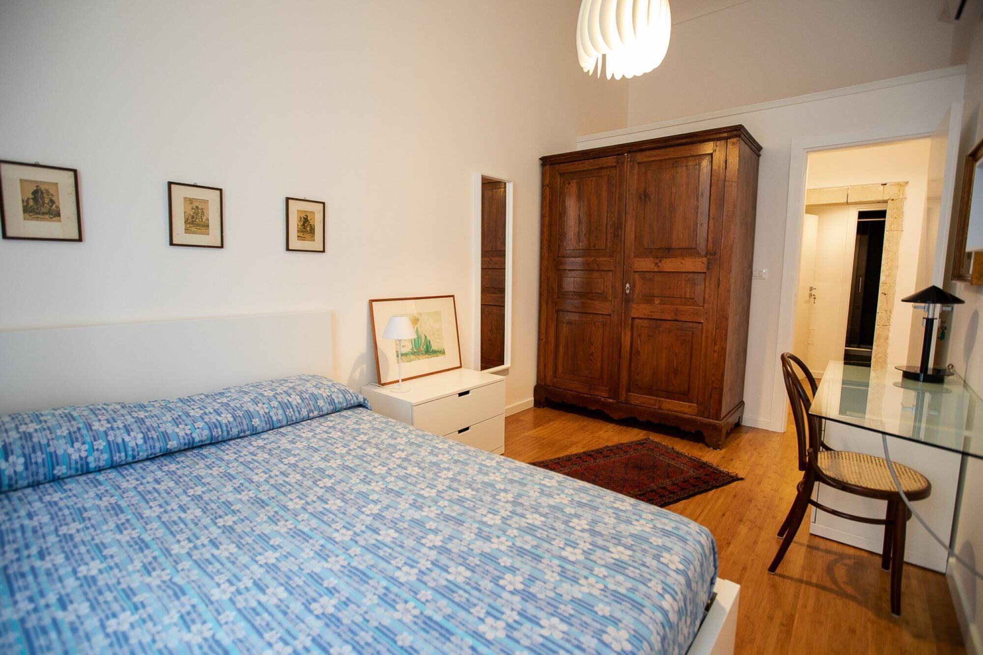 A bedroom with a bed, a wooden wardrobe, a desk with a chair, and framed pictures on the wall.