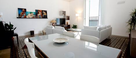 A modern living room with a dining table, white sofas, a TV, and artwork on the wall.