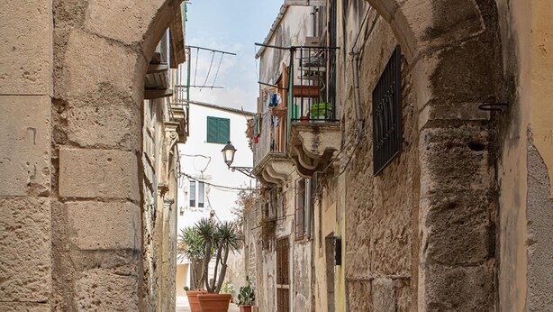 A narrow alleyway with stone walls and a tiled floor, leading to a building with balconies and potted plants. - Studio Dione 130 by Wonderful Italy (Syracuse)