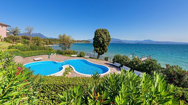 A swimming pool with a curved design, surrounded by lush greenery and a view of the sea. - San Sivino 55 con Piscina by Wonderful Italy (Manerba del Garda)
