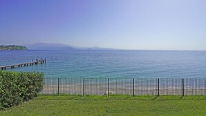 A coastal view with a wooden pier extending into calm waters, a grassy area in the foreground, and a distant mountain range under a clear sky. - San Sivino 55 con Piscina by Wonderful Italy (Manerba del Garda)