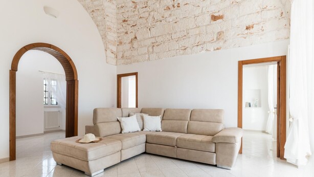 A spacious living room with a beige sectional sofa, a wooden archway, and a white tiled floor. - Villa Tranquillo 1876 by Wonderful Italy (Ostuni)