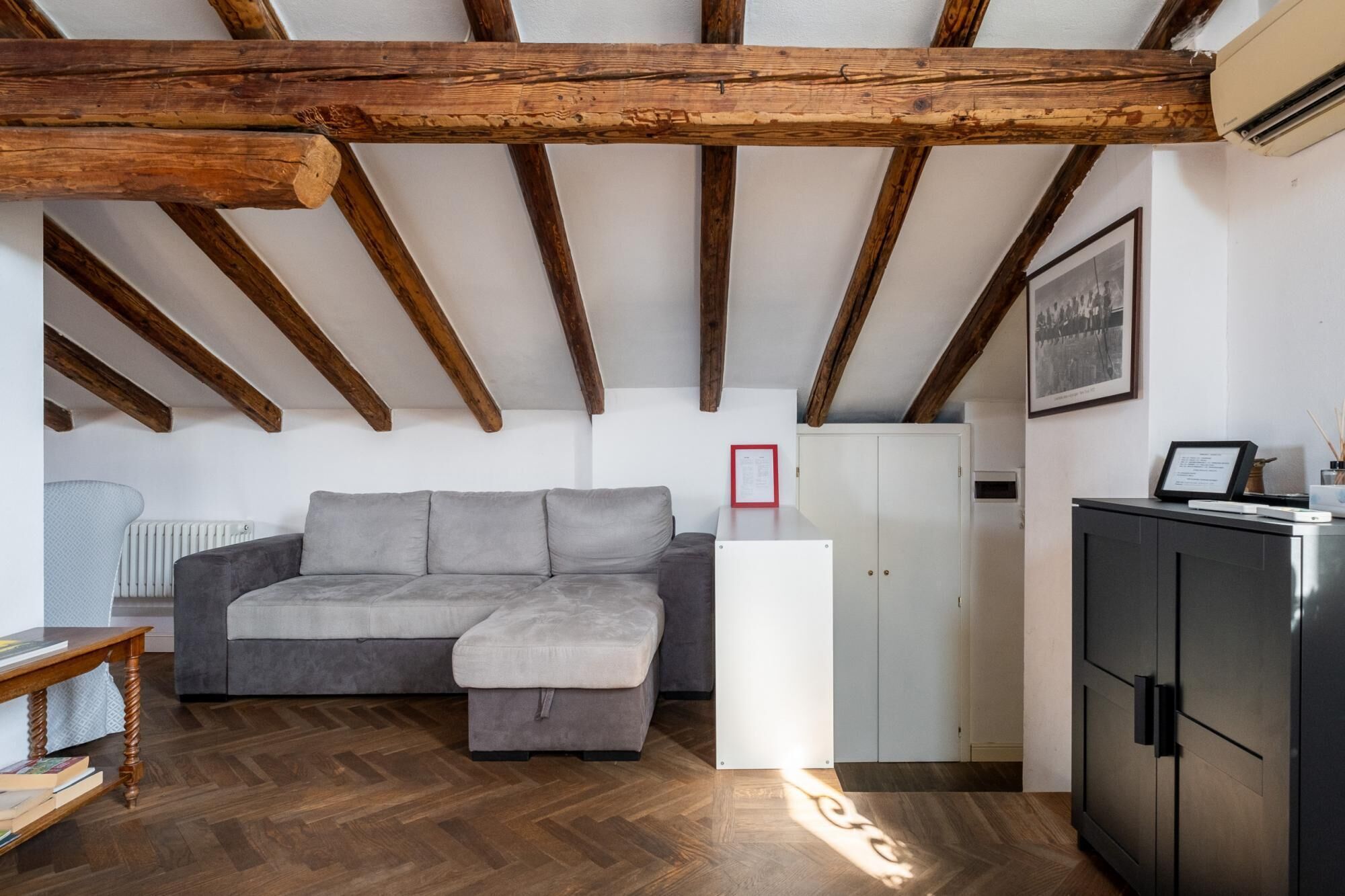 A loft-style room with exposed wooden beams, a grey sectional sofa, a white cabinet, and a radiator.