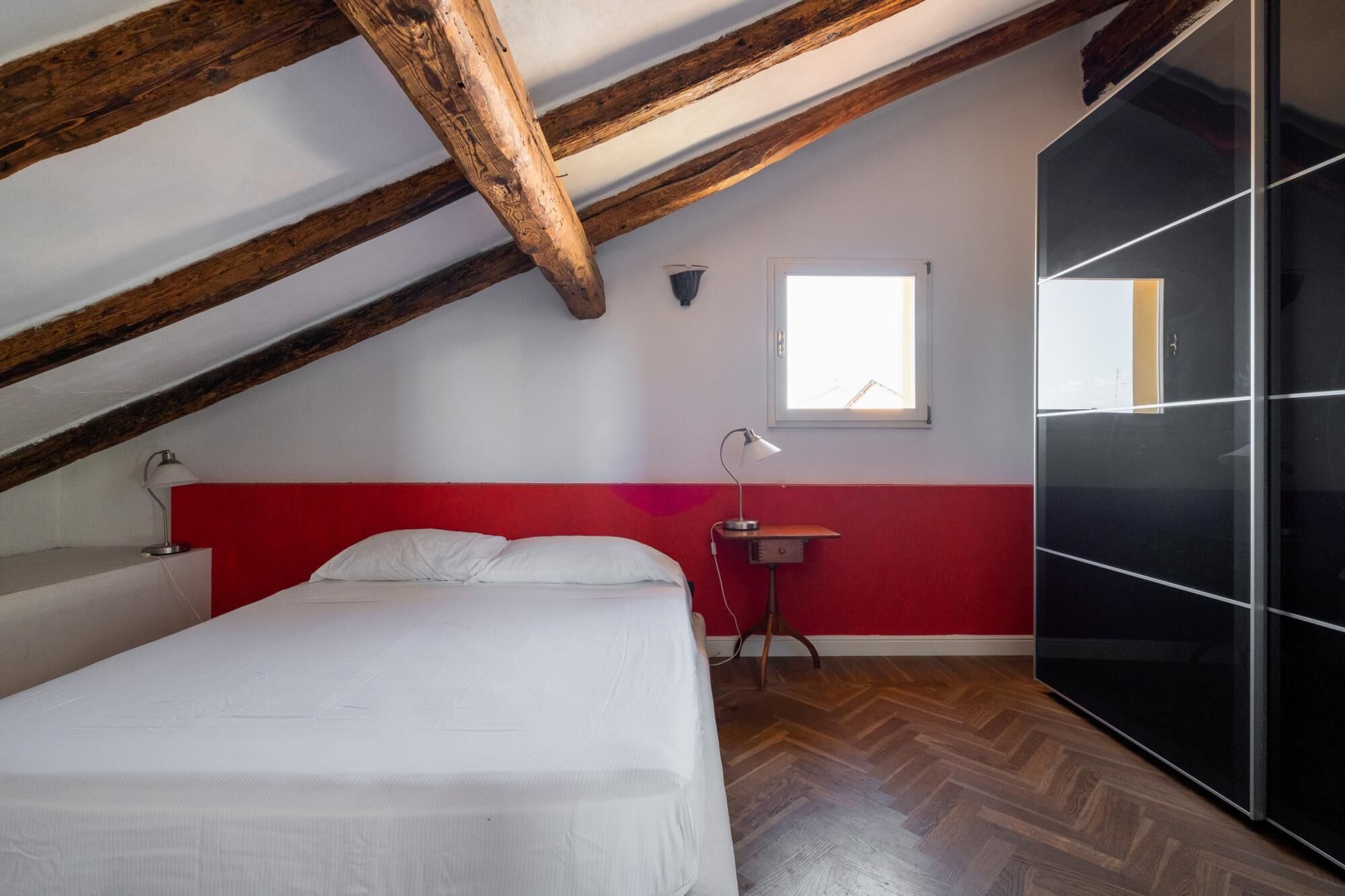 A bedroom with a sloped ceiling, a bed, a bedside table, a lamp, a window, and a wardrobe.