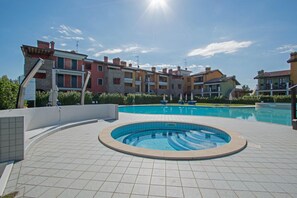 A swimming pool area with tiled flooring, a circular hot tub, and a row of colorful apartment buildings in the background. - Baglio Degli Ulivi 3 by Wonderful Italy (Sirmione)