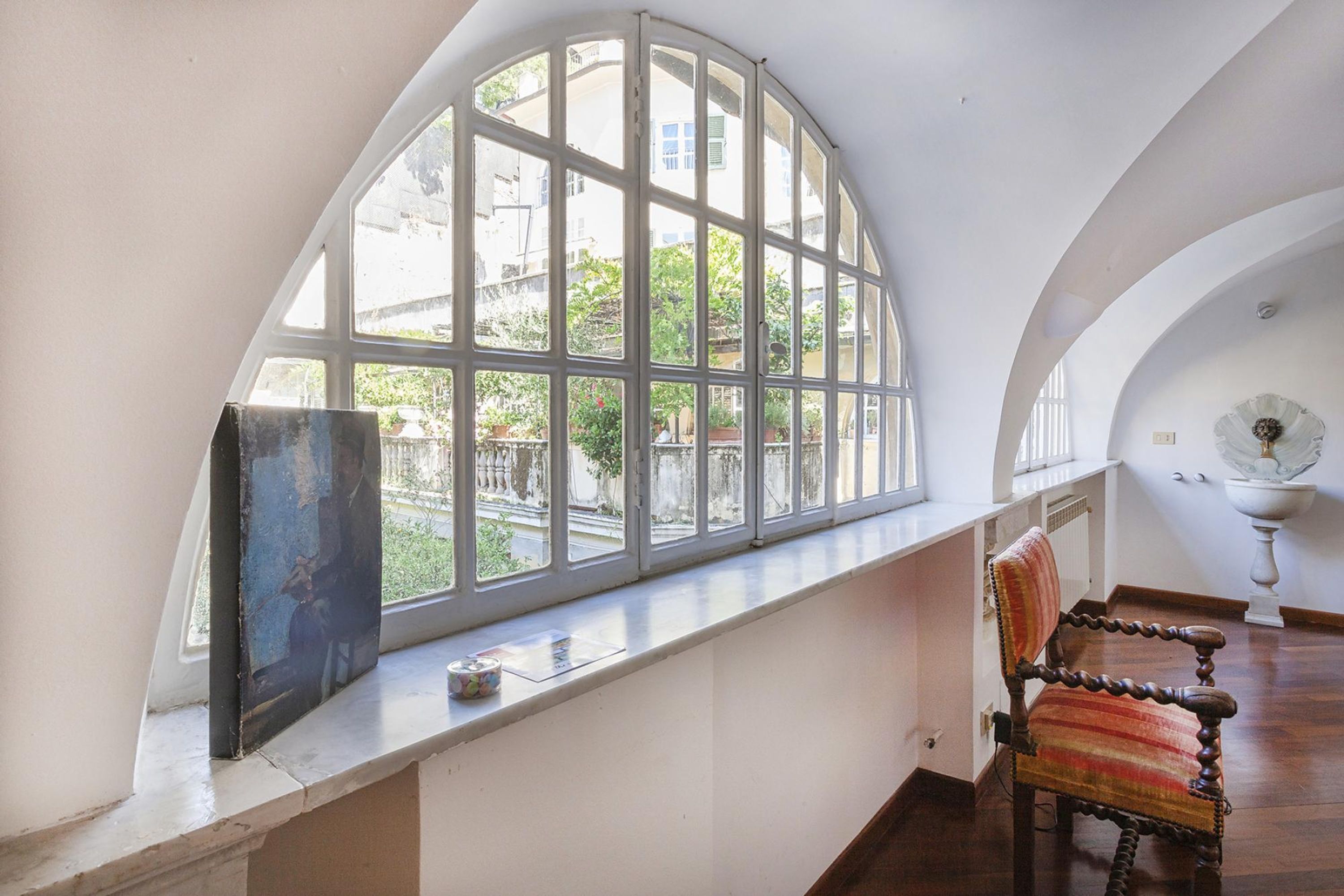 A room with a large arched window, a wooden chair, and a white pedestal lamp.