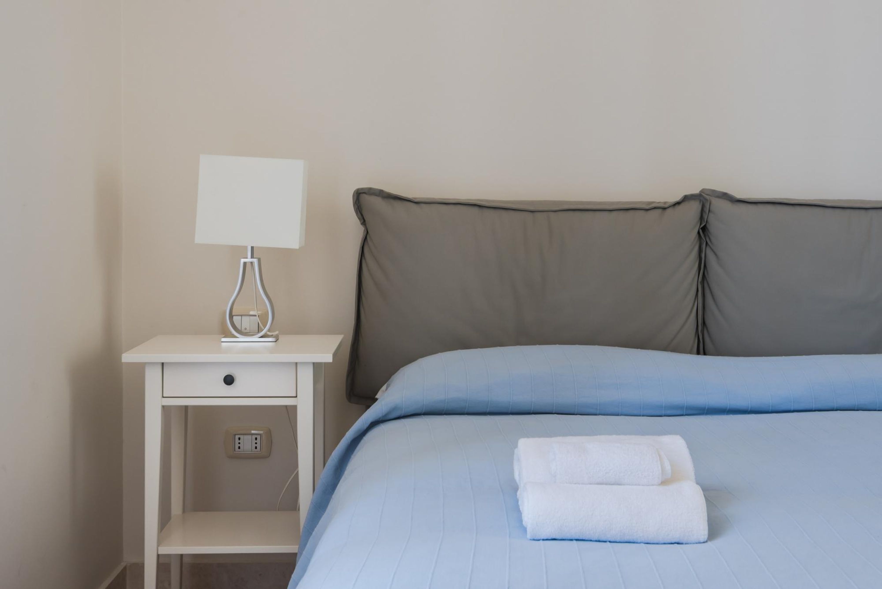 Een net opgemaakt bed met een blauwe sprei en twee kussens, een witte nachtkastje met een lamp, en een opgevouwen witte handdoek op het bed.