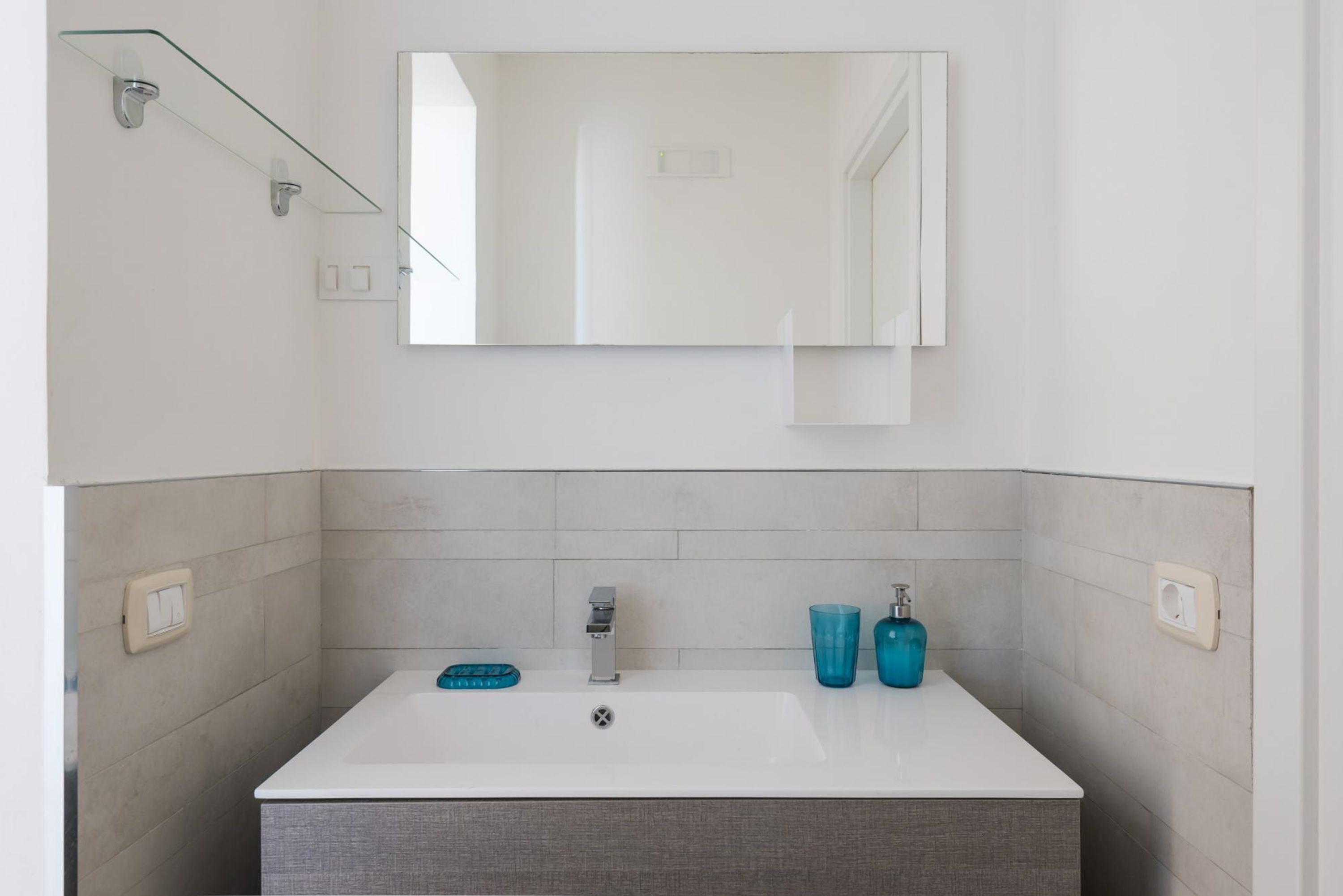 A bathroom with a white sink, a mirror, and a glass shelf.