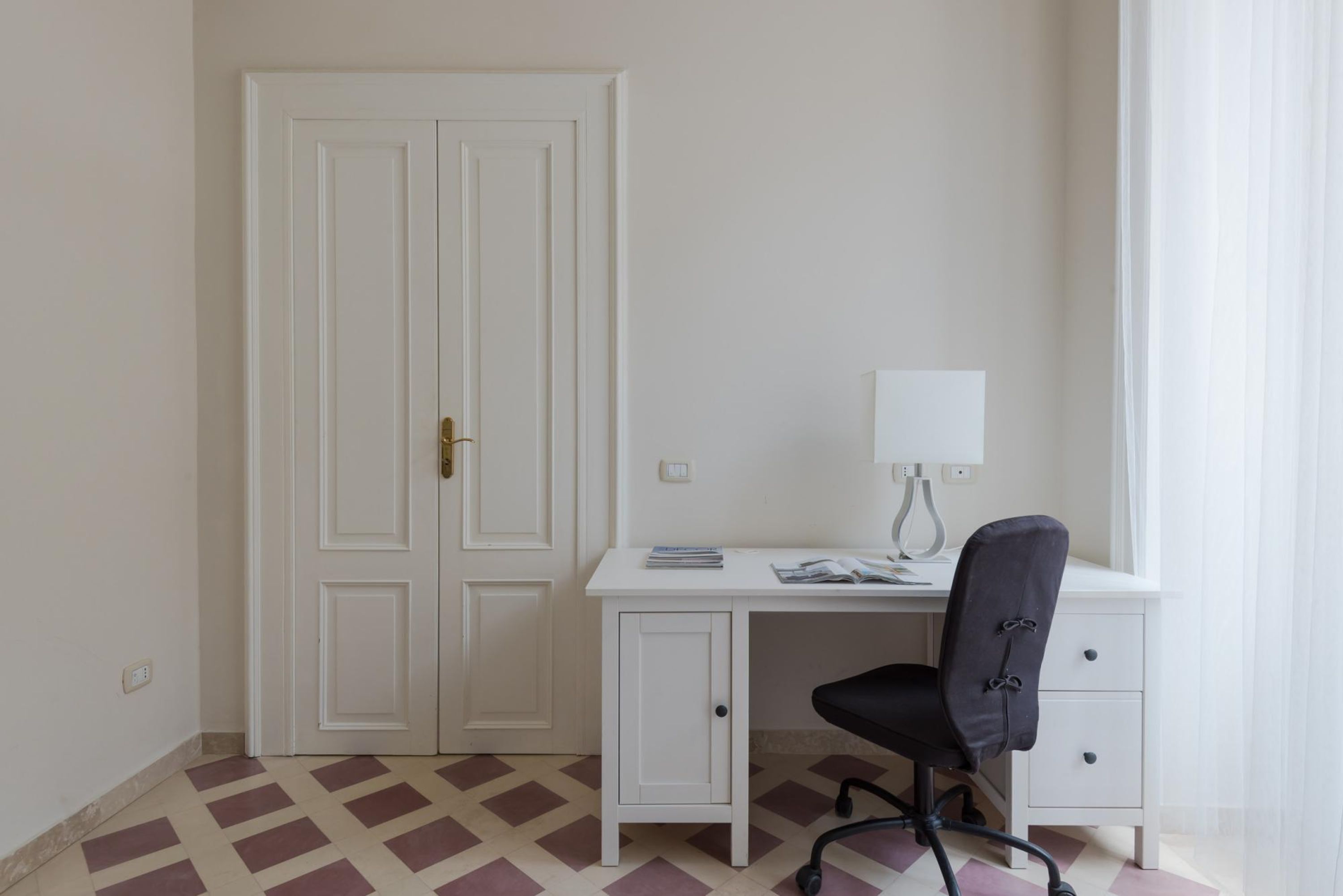 Ein Zimmer mit weißem Schreibtisch, schwarzem Bürostuhl, geschlossener weißer Tür und einem gemusterten Boden.
