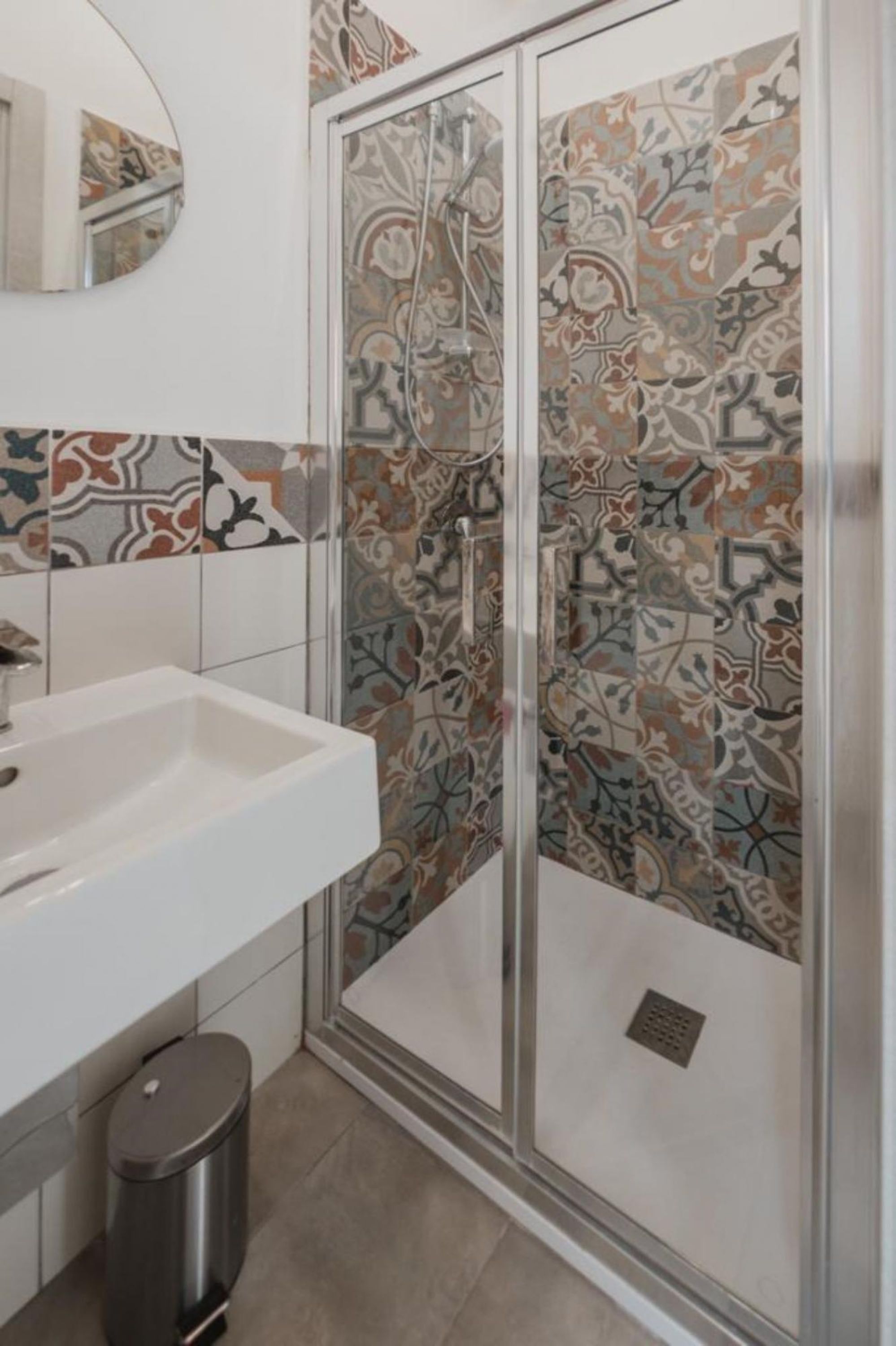 A bathroom with a white sink, a mirror, a shower with patterned tiles, and a trash bin.