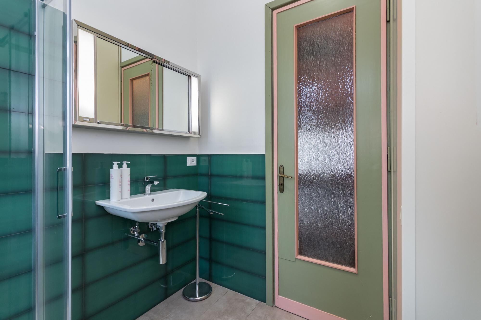 A bathroom with a glass shower enclosure, a white sink, and a green tiled wall.