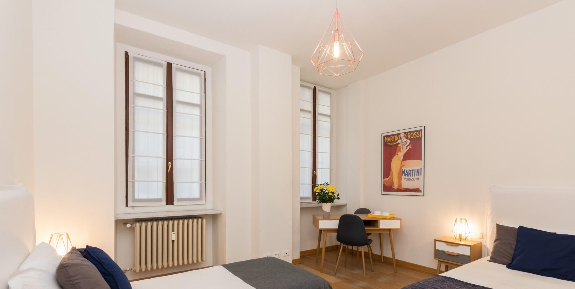 Une chambre d’hôtel moderne avec un lit, un bureau et une chaise. On y trouve un radiateur sous la fenêtre, une affiche au mur et un luminaire suspendu au plafond.
