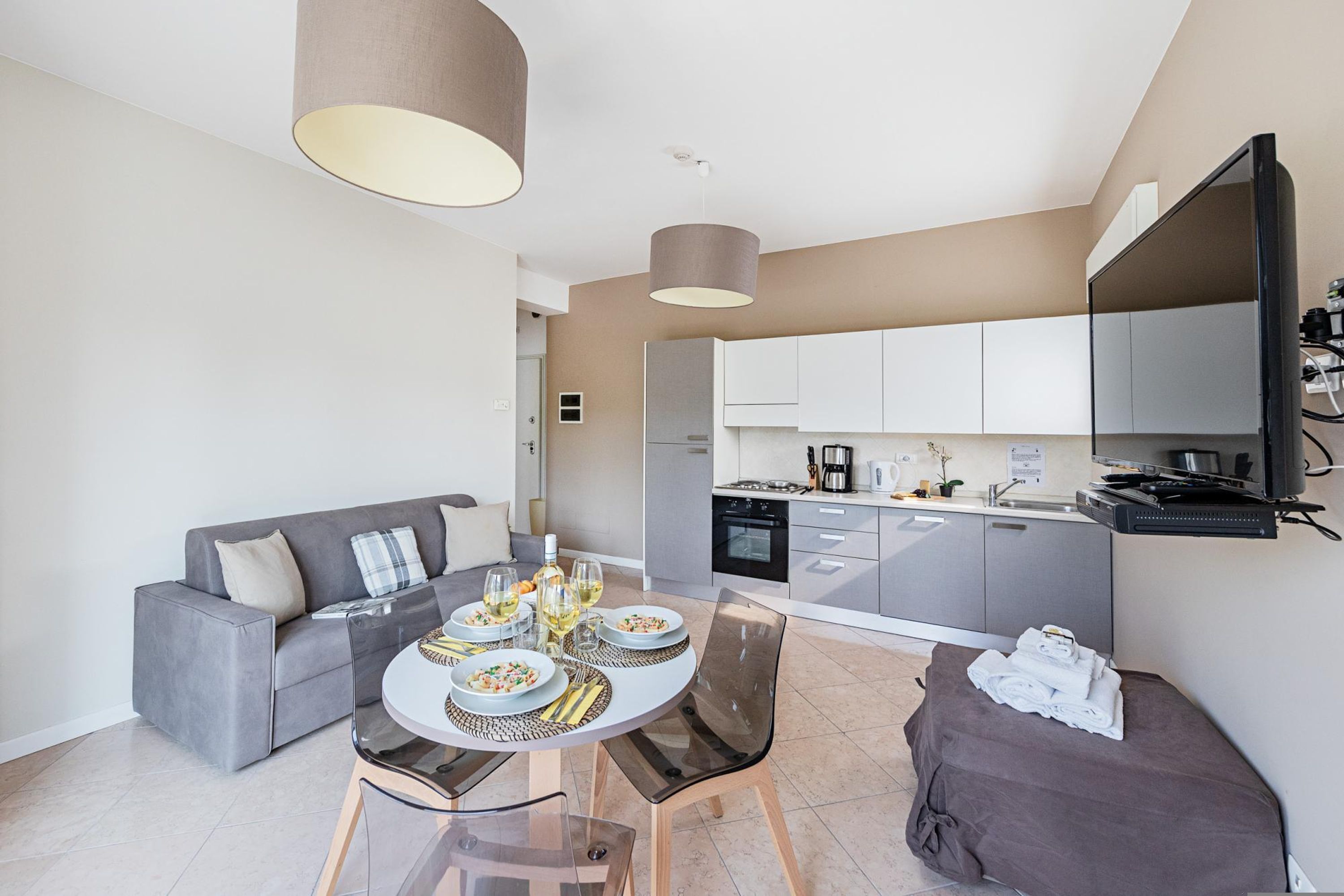 A modern living room with a grey sofa, a glass dining table set with plates and glasses, and a kitchen area with grey cabinets and a flat-screen TV.