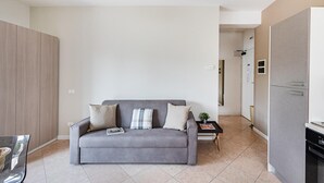A modern living room with a grey sofa, a small table, and a kitchen area with a fridge and oven. - Residenza Miralago - 10 Lavinia - Locazione BK (Desenzano del Garda)