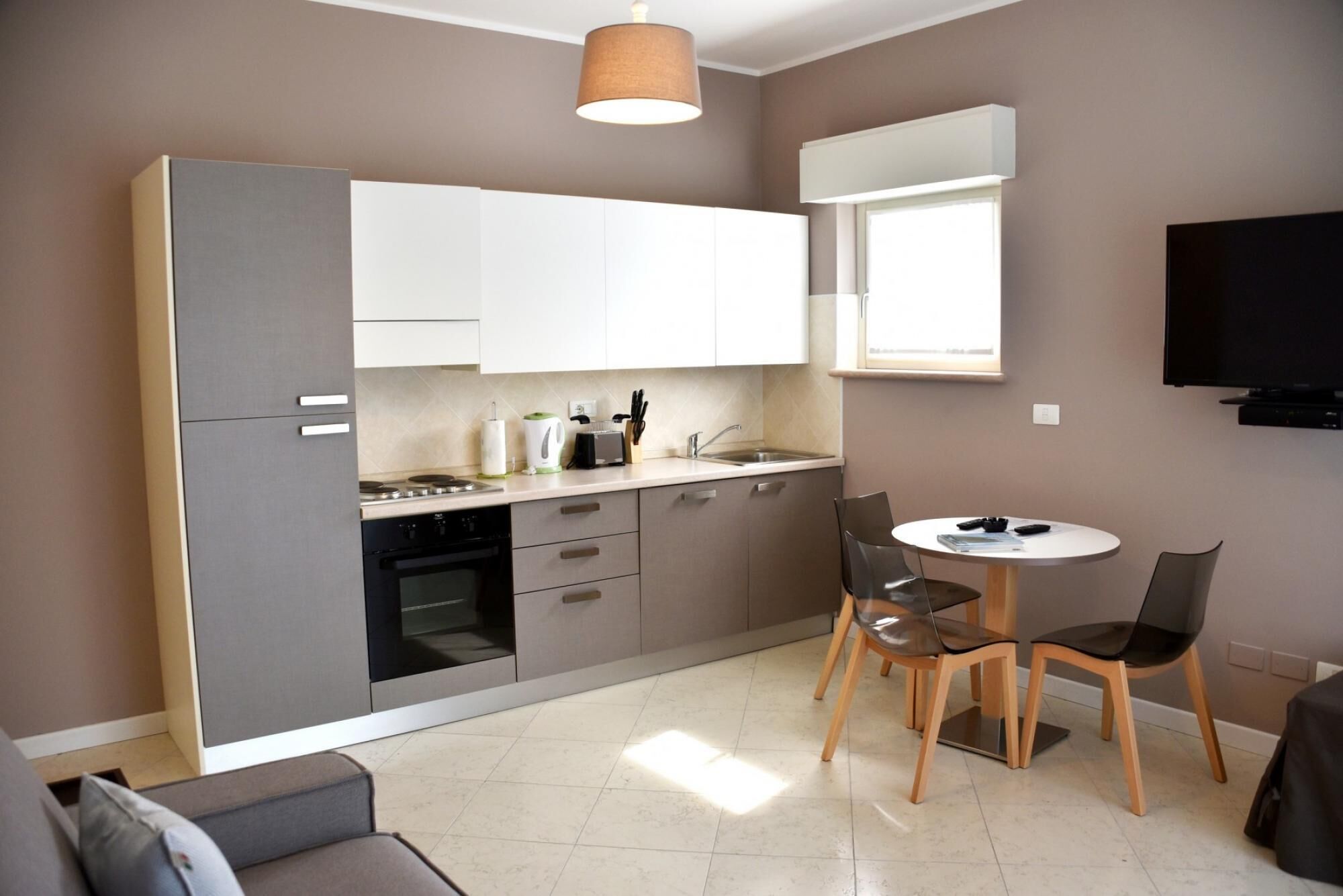 A compact kitchen with a dining area, a flat-screen TV mounted on the wall, and a window providing natural light.
