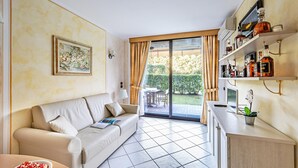 A modern living room with a sofa, a dining table with a plate of food, a kitchen area with shelves of bottles, and a sliding glass door leading to a garden.