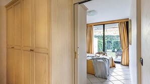 A room with a large wooden wardrobe, a bed with a grey blanket, and a view of a garden through a sliding door.