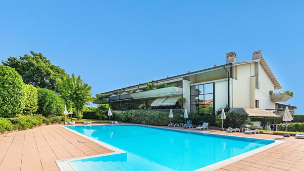 A modern building with a swimming pool, surrounded by greenery and sun loungers.