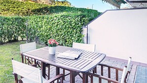 An outdoor patio with a wooden table and chairs, a hanging basket, and a view of a hedge and tree.