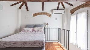 Un dormitorio con cama, pared de ladrillo curva, unidad de aire acondicionado, ventana con cortinas y suelo de madera.