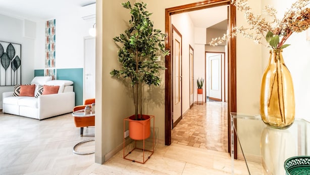 Ein modernes Wohnzimmer mit einem weißen Sofa, einem orangefarbenen Sessel und einem Blumentopf. Ein Flur führt in einen anderen Raum mit einem Spiegel und einer Blumenvase.