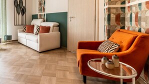 Ein Wohnzimmer mit einem weißen Sofa, einem orangefarbenen Sessel und einem Glastisch. Im Hintergrund ist eine Tür und eine Wand mit Muster zu sehen.