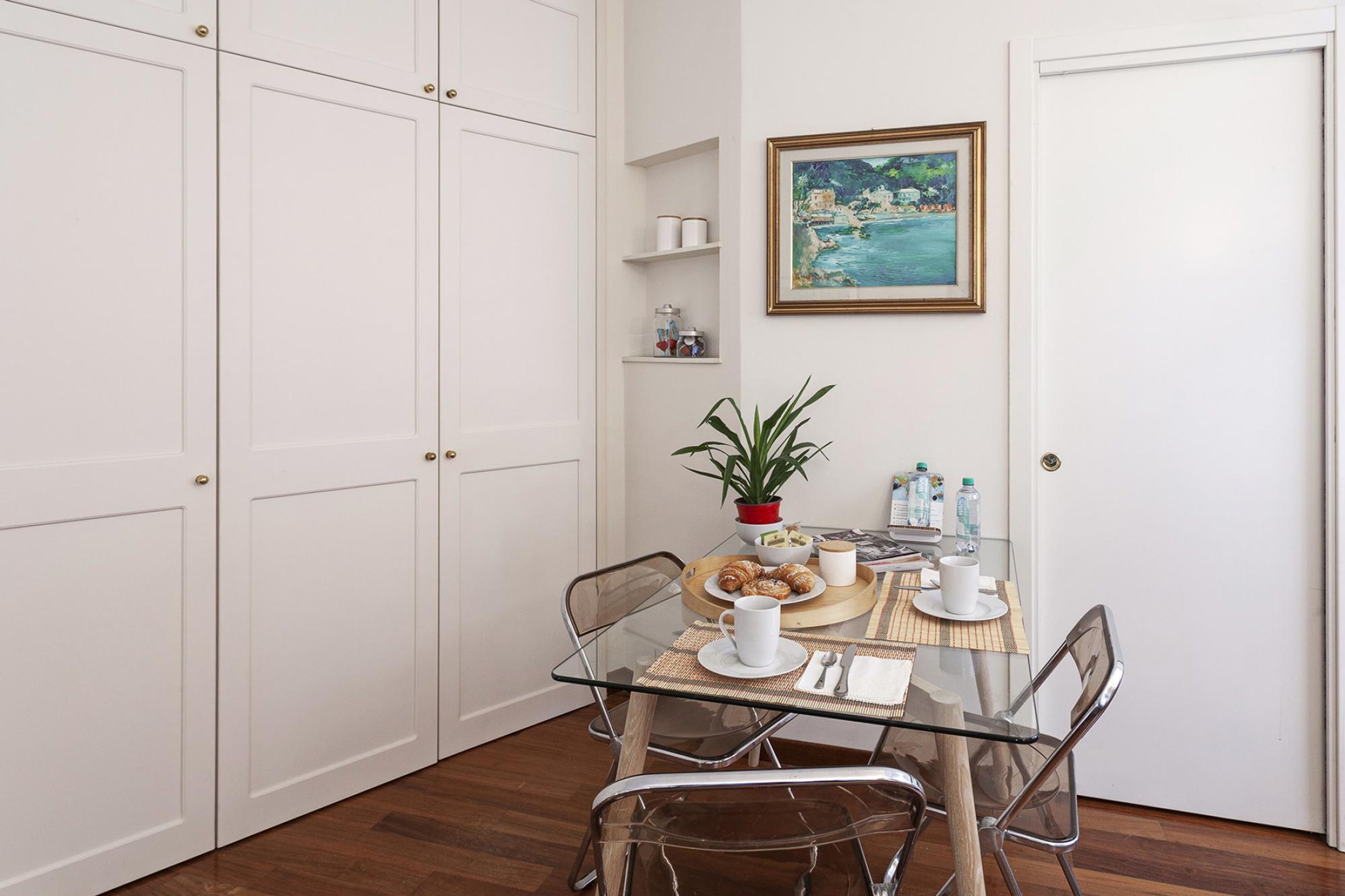Un'area pranzo con un tavolo in vetro, sedie, una pianta in vaso e un quadro appeso al muro.
