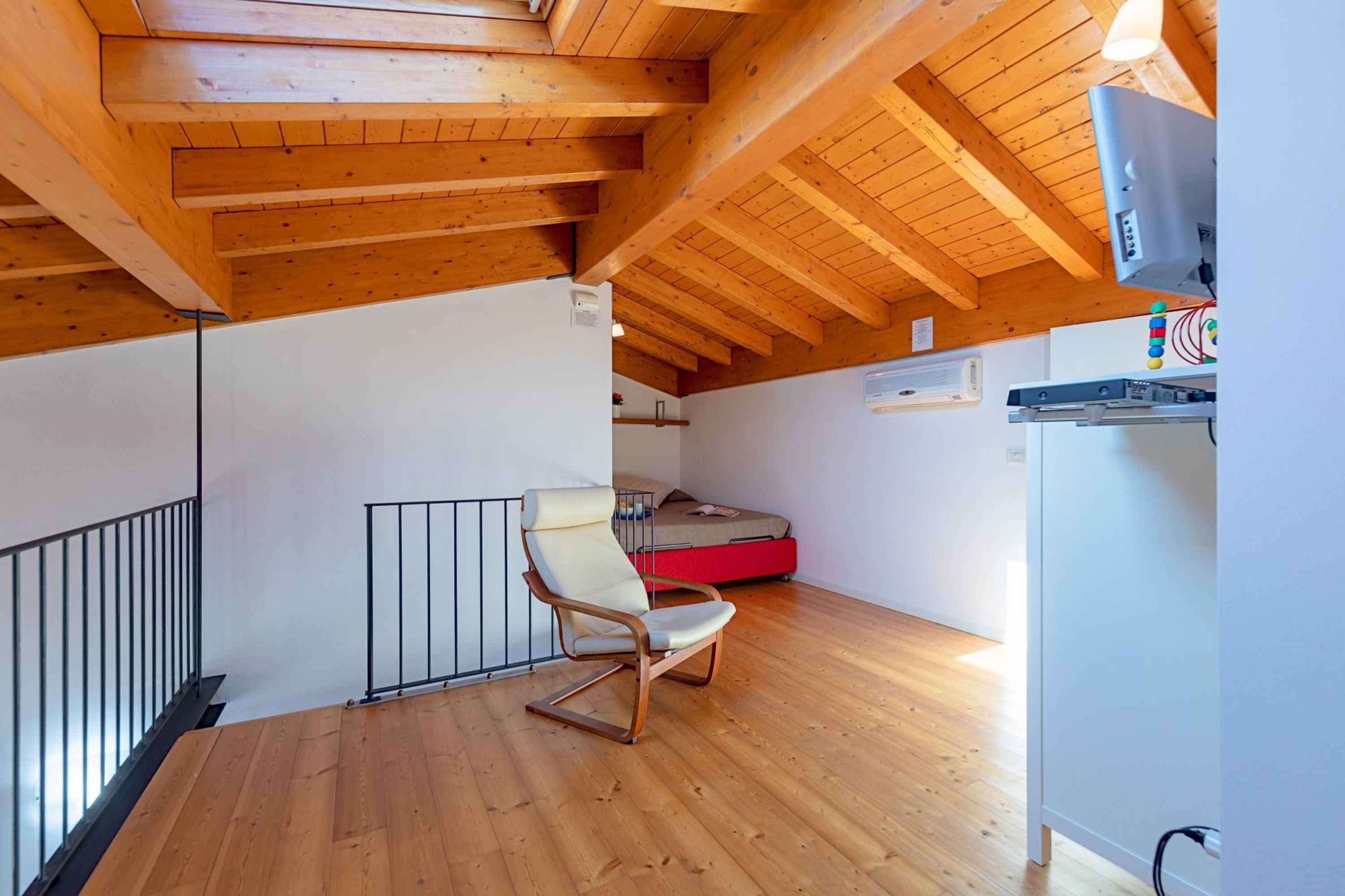 A room with a sloped ceiling, wooden beams, a single chair, a bed with a red mattress, and a small table with a television.
