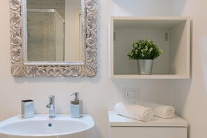 A bathroom with a white sink, two faucets, a mirror with an ornate frame, a shower, and a shelf with a potted plant. - Lake Sunbeam Villa by Wonderful Italy (Manerba del Garda)