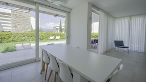 Ett modernt matområde med ett vitt bord och stolar, stora fönster och utsikt över grönska och en byggnad utanför.