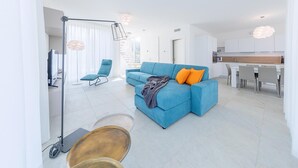 Ett modernt vardagsrum med en blå soffa, en golvlampa och en matplats, med ett kök synligt i bakgrunden.