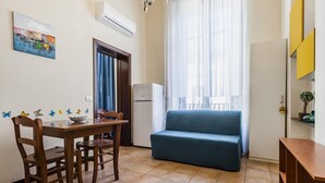 A small living area with a dining table, chairs, a sofa, a window with curtains, a wall-mounted air conditioner, and a colorful painting. - Grimaldi 8 Apartment by Wonderful Italy (Catania)
