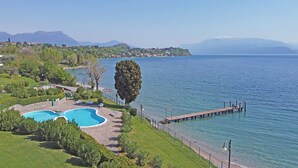A swimming pool with a stone deck, surrounded by greenery and a wooden dock extending into a lake. - San Sivino 42 con Piscina by Wonderful Italy (Manerba del Garda)