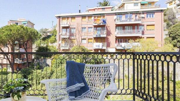 A balcony with a white wicker chair and a small table, overlooking a row of colorful apartment buildings. - Shelley Apartments - Tree by Wonderful Italy (Genoa)