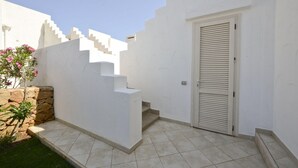 A white-walled building with a tiled patio, a stone wall, and a door with horizontal blinds.