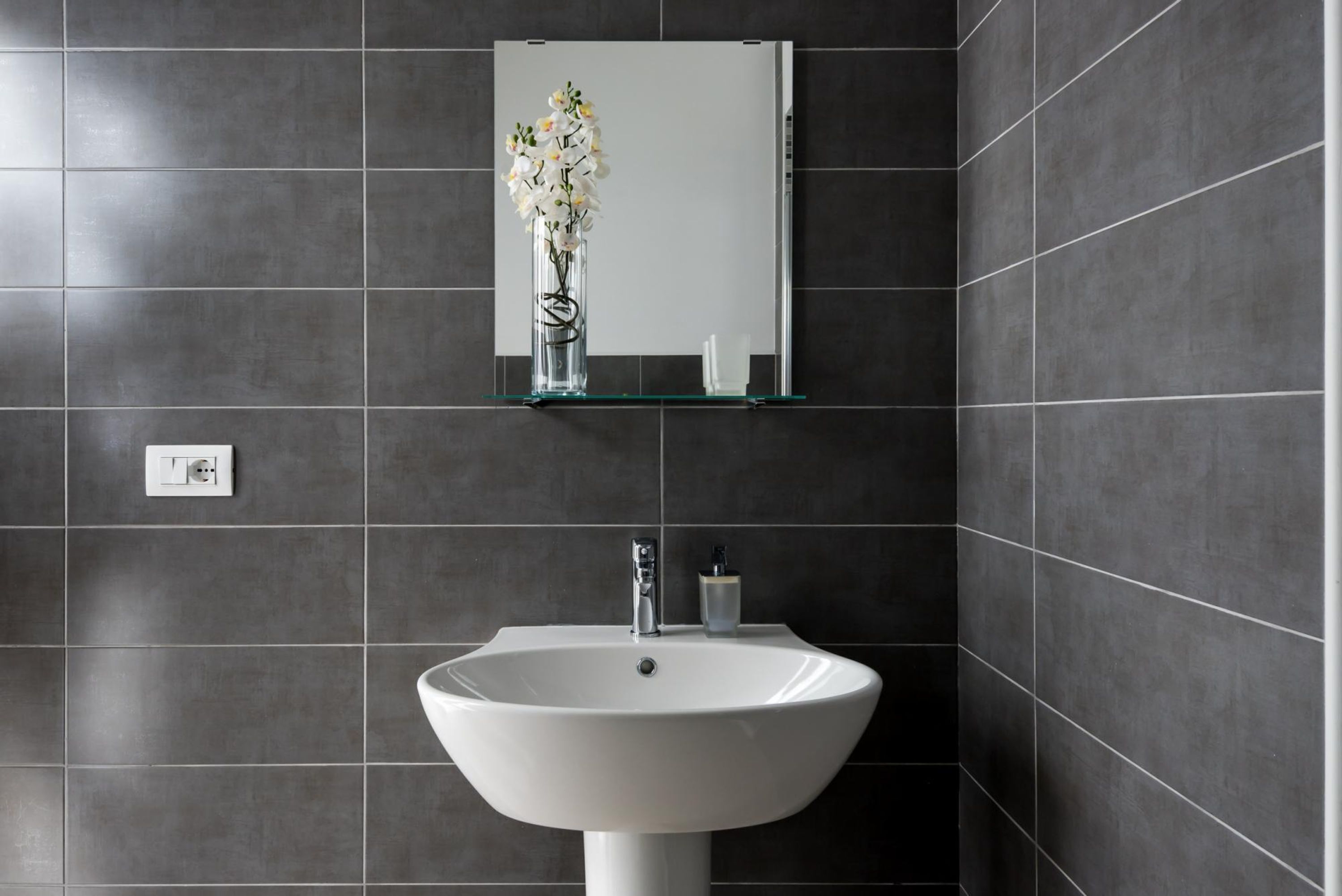 A bathroom with a white sink, a mirror above it, and a vase with flowers on a shelf.