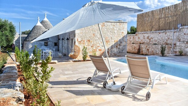 A poolside area with lounge chairs, a pergola, and a stone building in the background.
