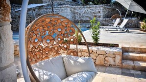 A hanging chair with a woven backrest and cushioned seat, a stone wall, and a pool area with lounge chairs.
