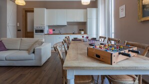 Ein gemütliches Wohnzimmer mit einer Couch, einem Esstisch, einem Tischfußballtisch und einer Küchenzeile mit weißen Schränken und einem Fenster.