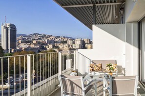 Un balcon avec vue sur la ville, des meubles blancs et une table agrémentée d’une plante.