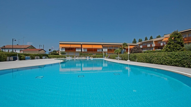 A swimming pool with a clear blue water surface, surrounded by a landscaped area with hedges and a building with orange and white exterior walls in the background. - Benacus E4 Apartment by Wonderful Italy (Sirmione)