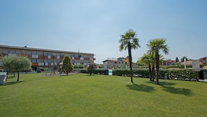 A courtyard with a lawn, palm trees, and a building with balconies. - Benacus E4 Apartment by Wonderful Italy (Sirmione)