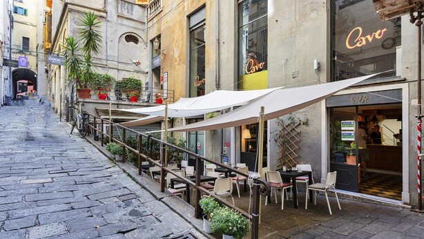 An outdoor café with tables and chairs, potted plants, and a building with a 'Cervo' sign. - Fancy Apartment in Palazzo Grimaldi by Wonderful Italy (Genoa)