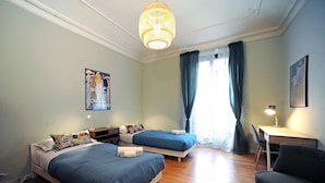 Une chambre avec deux lits, un bureau, une chaise, un luminaire suspendu et une fenêtre avec des rideaux.