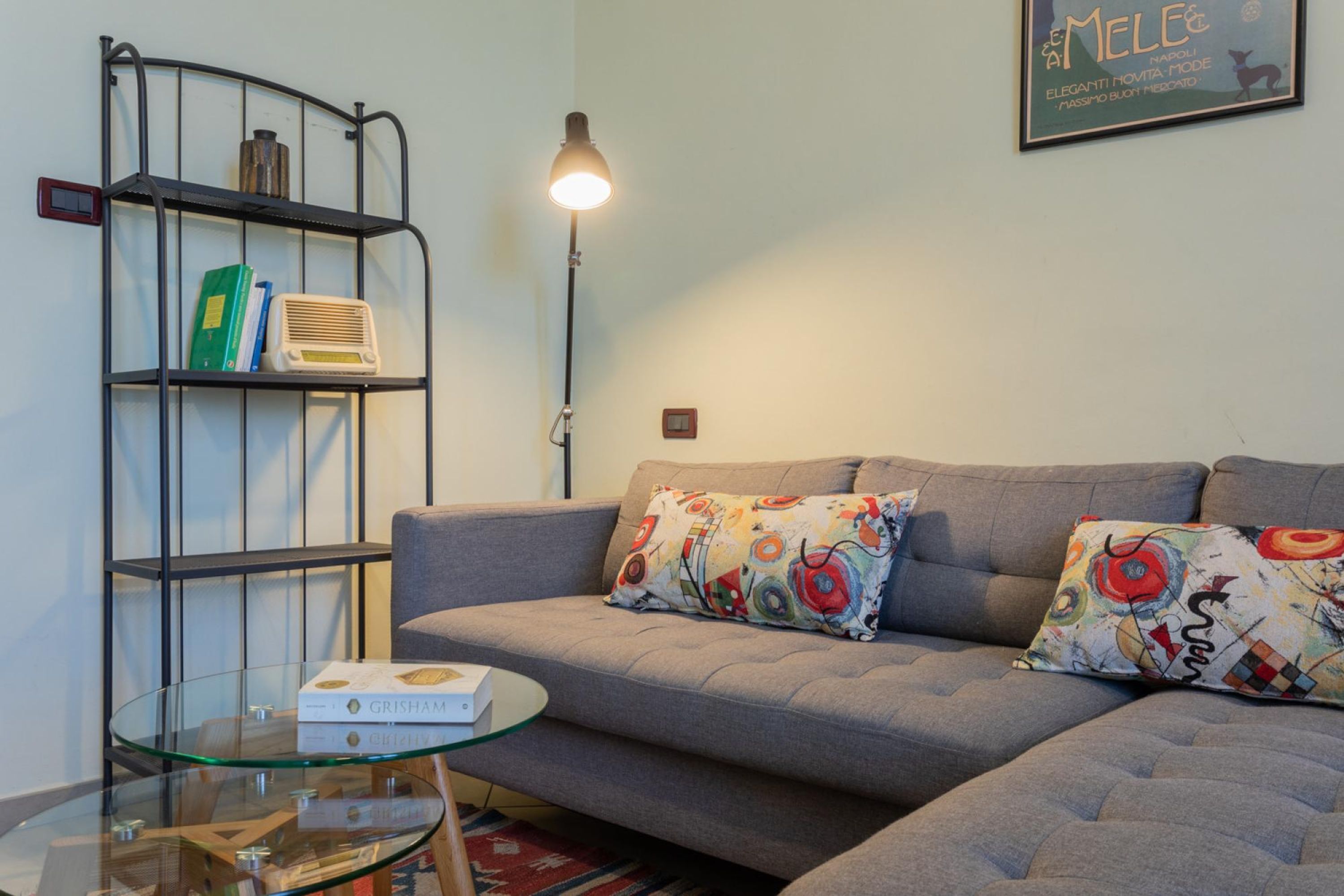 A living room with a grey sofa, colorful pillows, a glass coffee table, a metal bookshelf, and a floor lamp.