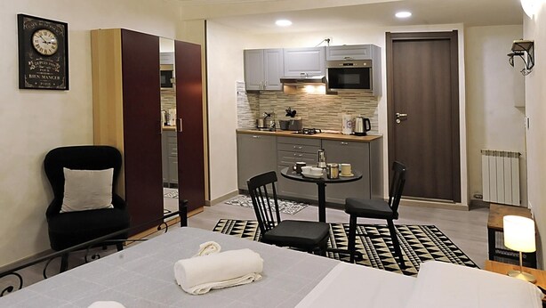 A compact hotel room with a bed, a small dining table, two chairs, a kitchenette, a clock, and a door.